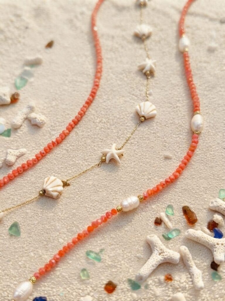 Collane estive color corallo ispirate al mare appoggiate sulla sabbia.